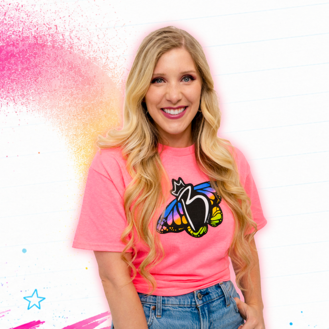 Brianna Playz wearing a bright pink graphic T-shirt with a black cat and colorful butterfly design, paired with blue jeans, against a white background with pink and gold paint splashes.