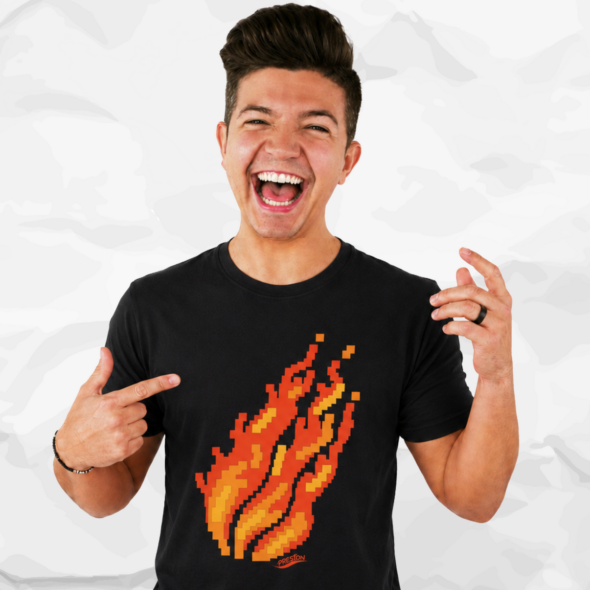 Preston Playz wearing a black crew-neck t-shirt with a pixelated orange and yellow flame graphic on the front, pointing at the shirt against a crumpled white paper background.